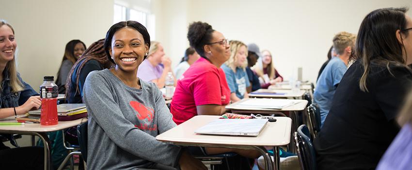 Female student smiling sitting at desk in classroom with other students.